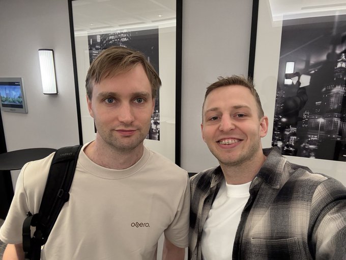 Two men pose for a selfie together indoors, one wearing a beige t-shirt with 'axera' text and a black backpack, the other in a plaid flannel shirt, with framed black-and-white photographs visible on the wall behind them.