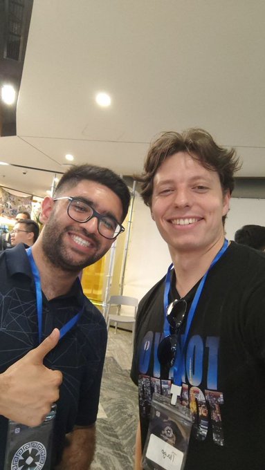 The image shows two men standing next to each other, both smiling for a photo. They are wearing t-shirts with the word "Solana" printed on them. The background of the picture appears to be a room or an event space, as there is a chair and a dining table visible in the scene. The post that accompanies this image reads: "Solana bros found each other at a monad event @xTiagoSantana".