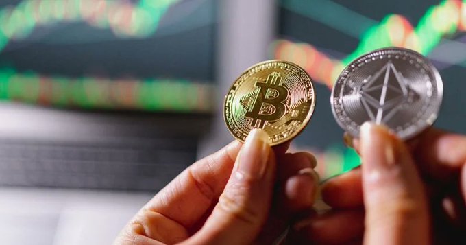 Two hands hold up physical Bitcoin and Ethereum coins against a backdrop of blurred green stock market charts.