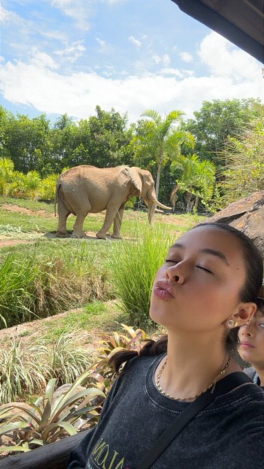 A selfie of a woman with closed eyes posing in front of an elephant at a safari or zoo, with lush greenery and another person visible in the background.