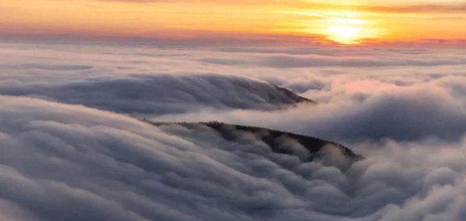 An aerial view captures a mountain peak rising above a sea of rolling clouds during a golden sunrise or sunset.