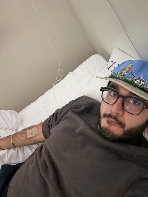 A bearded man wearing glasses, a colorful patterned cap, and a dark gray hoodie lies in a hospital bed with an IV visible on his arm.