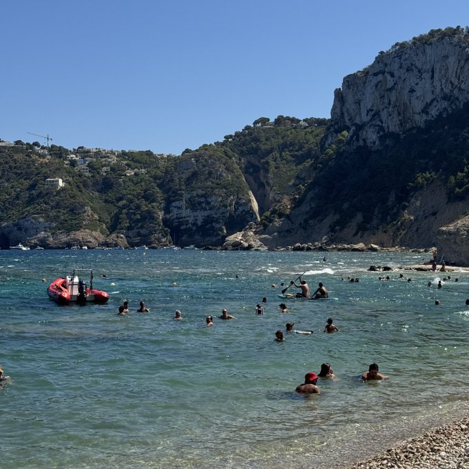A crowded beach scene with people swimming in turquoise waters near a rocky cliff, featuring a small red boat and a pebbly shore in the foreground—the person is using this scenic vacation photo to test if their post is visible after being shadowbanned, with the picturesque getaway highlighting their return to social media rather than commenting on any crypto development.