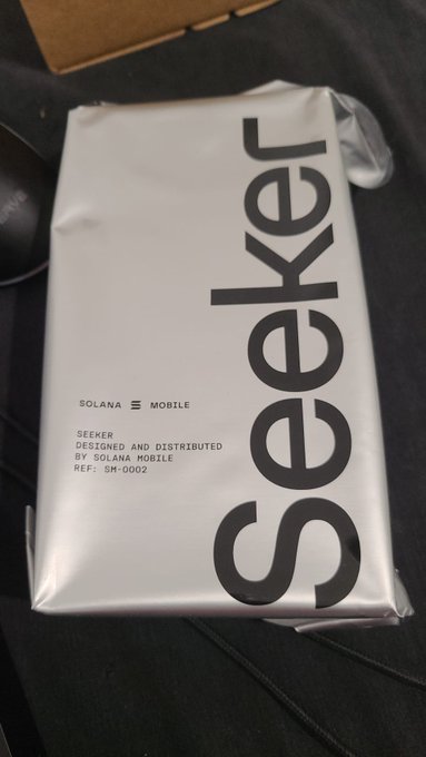 The image features a silver-colored box with black text that reads "Seker" on it, along with some other text. The box appears to be a phone case or packaging for a product named "Seeker", possibly related to Solana mobile. The context of the image suggests that someone has just received this item and is excited about exploring its features and testing out mobile dApps.