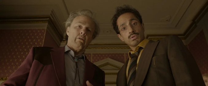 Two men in suits, one older with grey hair and a younger man with dark hair and a mustache, both looking down at the camera with serious expressions.