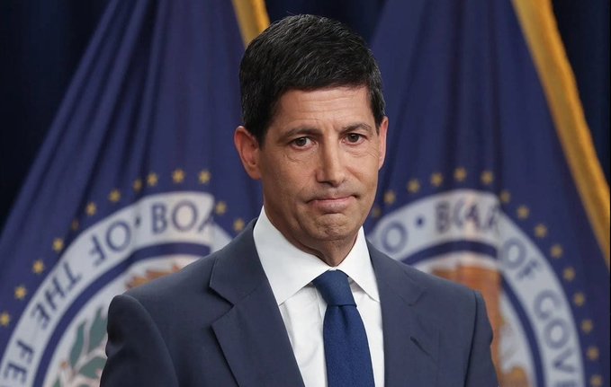 A man in a navy blue suit and blue tie stands before blue flags bearing the seal of the 'BOARD OF GOVERNORS OF THE FEDERAL RESERVE SYSTEM'.