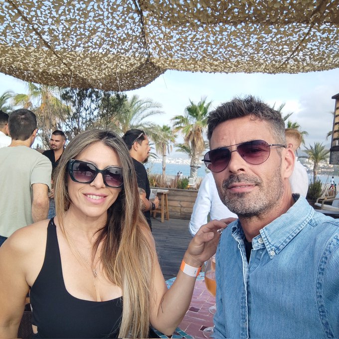The image shows a man wearing sunglasses, a woman with long blonde hair, and another person sitting at a table under an outdoor canopy. The people are gathered around a dining table, which has wine glasses placed on it. A group of people is also present in the background, suggesting that they might be attending an event or social gathering. The post about the @cryptocom event in Malaga seems to be related to this scene as it provides context for the location and the atmosphere at the event.