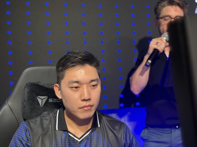 A focused young esports player wearing a "Secretlab" branded jersey sits intently in a gaming chair with a blue polka-dot pattern backdrop, while a bearded commentator holding a microphone stands behind him, perfectly capturing the "insomniac locked in" competitive gaming atmosphere.