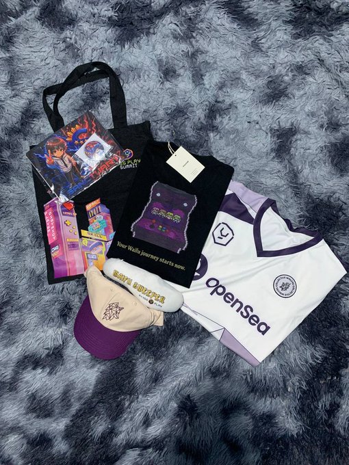 A collection of merchandise gifts laid out on a gray fluffy rug, including a "Waifu Sweeper" item from @YGG_Play, a black "Yield Guild Summit" tote bag, an OpenSea jersey, and a WolvesDAO shirt and cap — matching the post about receiving these items from @YGG_Play, @aemacgg, @TitaKeith, and @WolvesDAO.