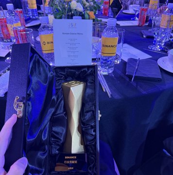 A hand holds an elegant black and gold trophy inscribed with "BINANCE" and Chinese characters, displayed in a presentation box at a formal awards banquet with bottled water and a menu visible on the table. The image aligns with the post's theme of recognizing Binance's contributions to the crypto community, serving as tangible visual evidence of an award honoring the company's achievements amid criticism.