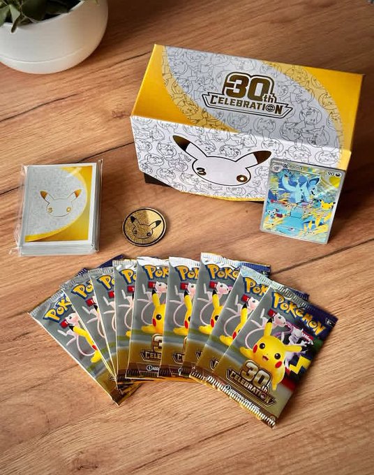 A Pokémon 30th celebration merchandise display on a wooden surface, featuring a yellow Pikachu-themed box, multiple Pokémon card booster packs, a coin, and a card sleeve with a Pikachu illustration.