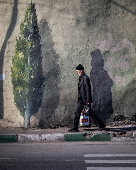 An older man in a black suit and cap walks along a sidewalk carrying a shopping bag, passing a weathered wall painted with a mural of a cypress tree.