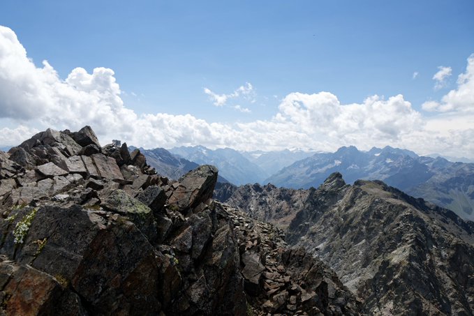 A mountain landscape featuring rocky peaks in the foreground with distant mountain ranges under a bright blue sky with scattered clouds.
