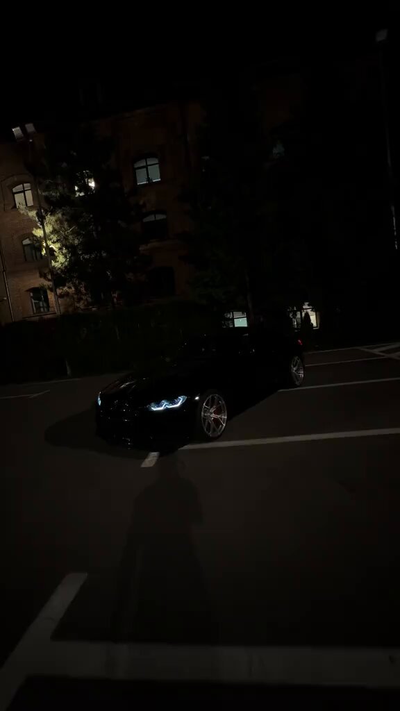 The image features a black car driving down a street at night. The car has blue headlights, which stand out against the darkness of the scene. In the background, there are buildings that add depth to the image. The overall atmosphere suggests an urban setting with nighttime activities.