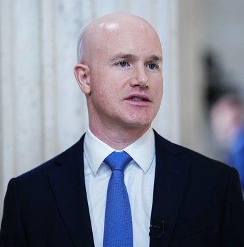 Brian Armstrong, CEO of Coinbase, is shown in a dark suit and blue tie during what appears to be an interview setting, with a small microphone clipped to his lapel. His serious expression corresponds with his recent bold prediction that Bitcoin could reach $400,000 by 2030.