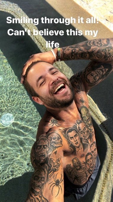 A heavily tattooed shirtless man smiles broadly while leaning back in a pool, with the text "Smiling through it all! Can't believe this my life" overlaid on the image.
