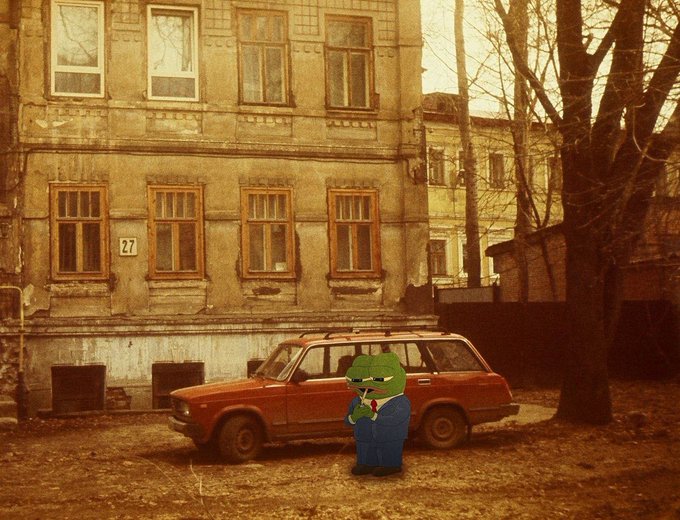 A vintage-style photograph of an old apartment building with a red car parked in front, edited to include a small cartoon frog in a blue suit smoking a cigar.