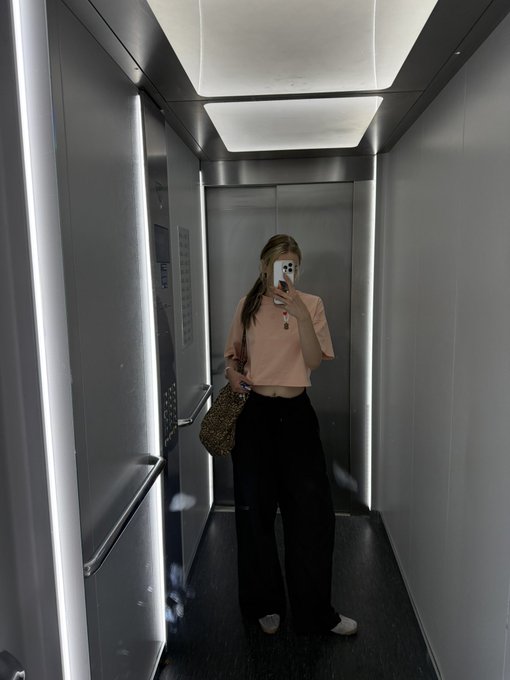 The image features a woman taking a selfie inside an elevator, holding her phone up to capture the moment. She is wearing a pink shirt and black pants. The background of the photo includes a gray wall with a light switch located above the elevator doors. There are also two handbags visible in the scene - one near the center-left side of the image and another closer to the bottom right corner.