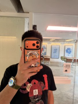 A person wearing a black graphic t-shirt and a wristwatch takes a mirror selfie with a smartphone featuring an orange patterned case, holding the phone in front of their face.