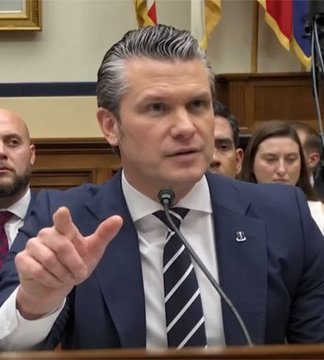 A man in a dark suit with a striped tie speaks at a congressional hearing, gesturing with his right hand while seated before a microphone.