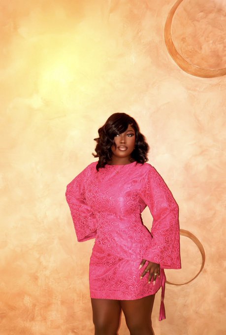 A studio portrait of a woman with shoulder-length dark hair wearing a vibrant pink, textured mini dress with long bell sleeves, posed against a warm golden backdrop.