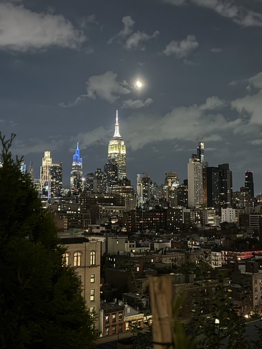 A nighttime cityscape of New York City featuring the Empire State Building illuminated under a full moon and cloudy sky.