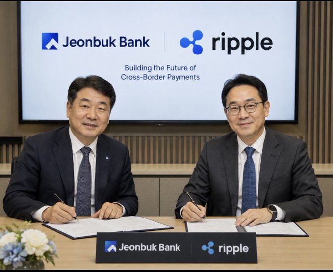 Two businessmen in suits sit at a table signing documents in front of a screen displaying 'Jeonbuk Bank' and 'ripple' logos with the text 'Building the Future of Cross-Border Payments'.