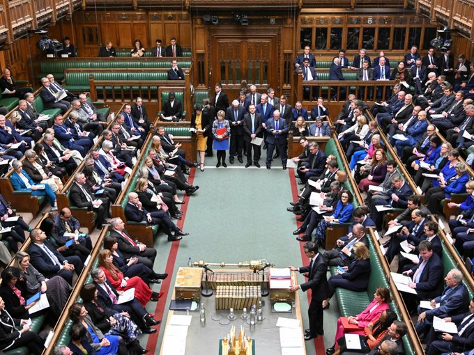 The interior of the UK House of Commons chamber with MPs seated on green benches facing each other, with officials standing in the center aisle.