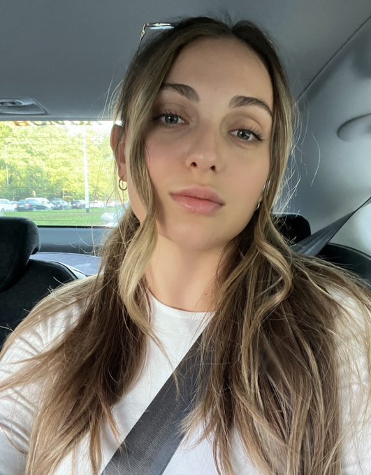 A young woman with long blonde hair sitting in a car, wearing a white t-shirt, with sunglasses perched on her head and a seatbelt across her chest.