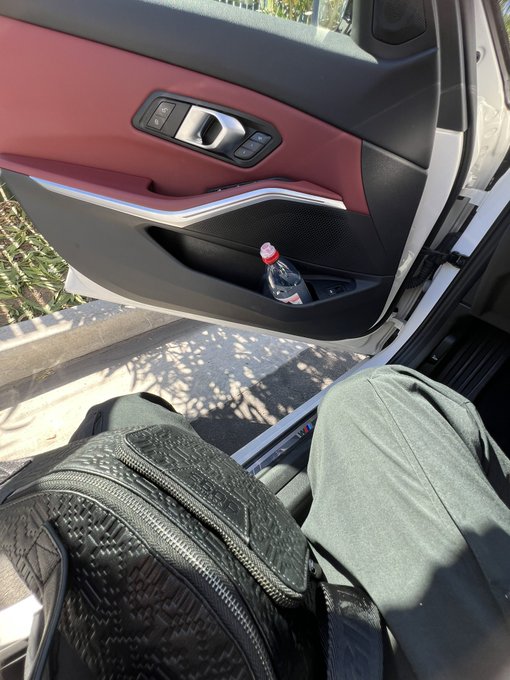 A photo taken from the passenger seat of a car with a red and black interior, showing a water bottle tucked into the door's storage pocket and a black backpack on the seat between the passenger's legs.