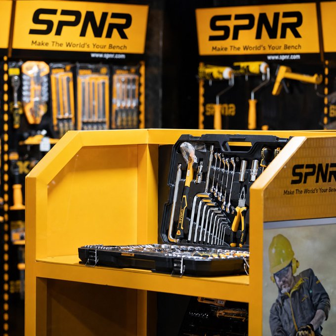 A display of SPNR brand tools, featuring wrenches and socket sets arranged on a bright yellow bench setup with branded signage reading 'Make The World's Your Bench'.