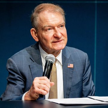 An older man in a blue plaid suit with an American flag lapel pin, speaking into a microphone at what appears to be a panel or speaking event.