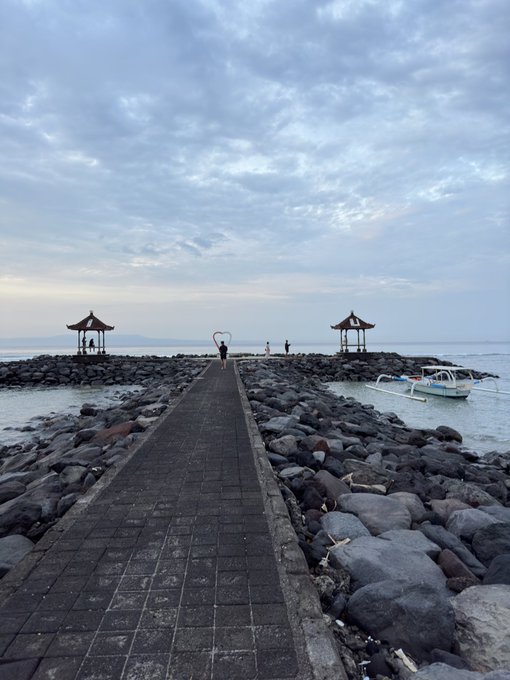 A stone-paved pier extending over a rocky breakwater into the sea, with two small pavilions at the end, a heart-shaped sculpture, and a boat anchored to the right under a cloudy sky.