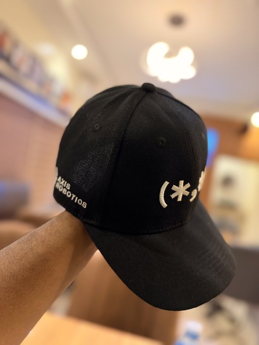 A black baseball cap featuring "Axis Robotics" branding on the side and a kaomoji-style logo on the front is held up, representing the collaboration merchandise from the Axis Robotics x Kaito epoch mentioned in the post.