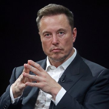 A close-up photograph of Elon Musk wearing a dark suit and white shirt, sitting against a black background with his hands clasped together in front of him.