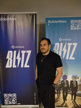 A man poses in front of "BuilderMare" and "Monad BLITZ" banners (featuring QR codes) at a Monad Blitz event, where he presented his project DAYAI—a decentralized AI model marketplace built on Monad that merges artificial intelligence with blockchain technology.