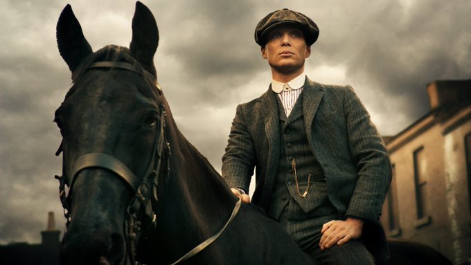 A man in a flat cap and grey three-piece suit sits on a black horse against a cloudy sky, in a scene resembling Peaky Blinders.