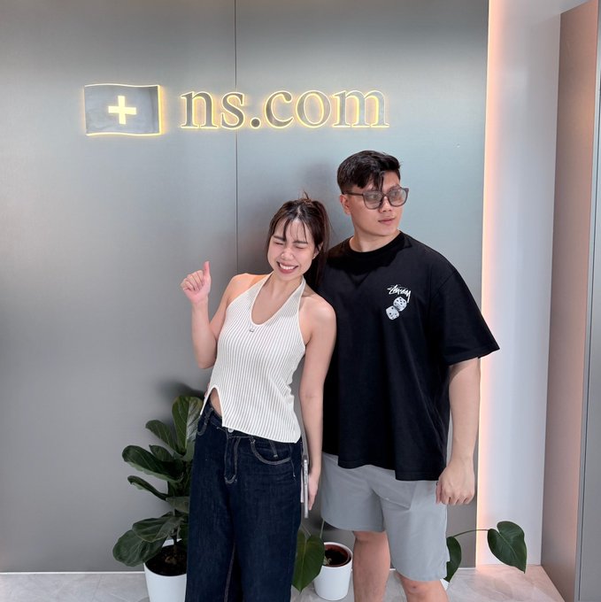 The image features two people, a man and a woman, standing next to each other near a wall with a sign that says "NS.COM" above them. The man has his arm around the woman's waist, while she stands beside him. They are both smiling at the camera, creating an endearing scene.

In front of them is a potted plant and several vases placed on a table or shelf behind them. There is also a keyboard visible in the background, suggesting that they might be in a room with some work-related items present.
