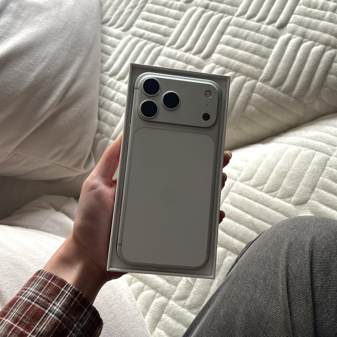 A hand holds an unboxed silver smartphone with a large triple-camera module on a white quilted bedspread.
