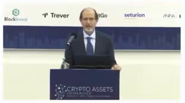 The image shows a man standing at a podium, giving a speech or presentation. The background of the image features a white wall with various logos and text, including "blockinnvest" and "Crypto Assets", suggesting that this event may be related to cryptocurrency or blockchain technology. The speaker is dressed in a suit and tie, indicating a formal setting for the event.