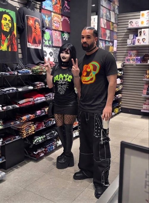 Two people posing in a t-shirt store; one wears a black Motley Crue shirt with fishnet tights and platforms, while the other sports a rasta-themed shirt and black cargo pants.