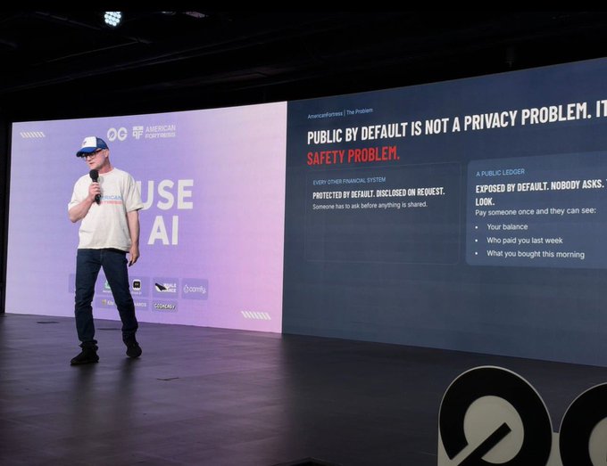 A man in a white t-shirt and baseball cap speaks into a microphone on stage. The screen behind him displays a presentation titled 'PUBLIC BY DEFAULT IS NOT A PRIVACY PROBLEM. IT IS A SAFETY PROBLEM.'