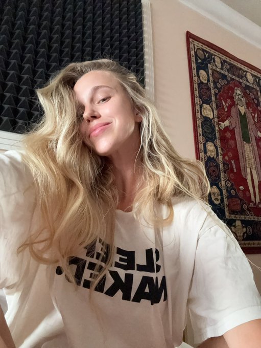 A selfie of a blonde woman wearing a white t-shirt with "MAKE [something] GREAT" text in a room with acoustic foam panels and an ornate tapestry on the wall.