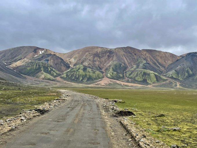 A landscape photograph of a dirt and gravel road leading toward rolling mountains with green vegetation and some snow patches under a cloudy gray sky.