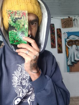 A person wearing a yellow beanie and blue hoodie holds up a green illustrated card to partially obscure their face, revealing one eye, a beard, and hand tattoos.