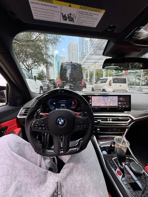 View from the driver's seat of a BMW, showing the steering wheel, dashboard display with navigation, and traffic including a bus ahead on a city street with tall buildings.