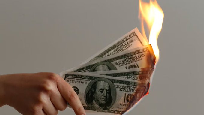 A hand holding a burning $100 bill engulfed in flames against a gray background.