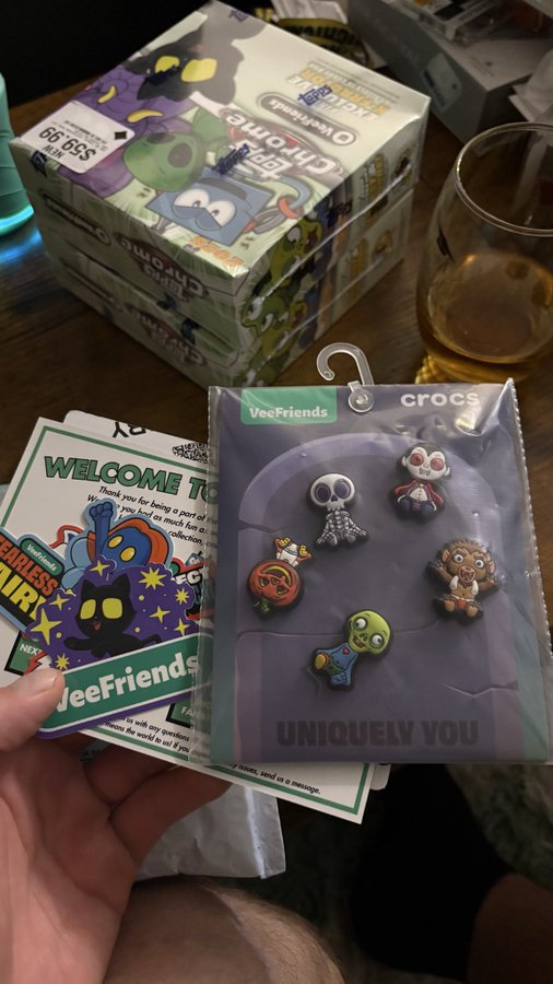 A hand holding a 'VeeFriends crocs UNIQUELY YOU' package of character charms, with 'Crocs Charlie' and 'Crocs Gerald' boxes stacked in the background alongside a welcome card.