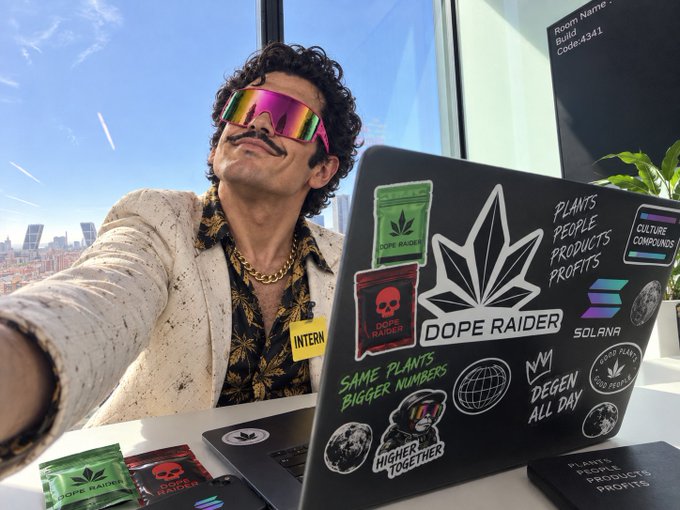 A man wearing iridescent sunglasses and a fake mustache poses for a selfie with a laptop covered in stickers including "DOPE RAIDER," "SOLANA," "CULTURE COMPOUND," and "DEGEN ALL DAY," along with branded product packets.
