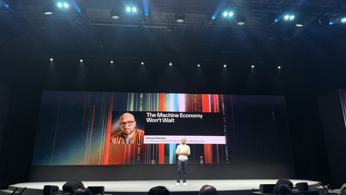 A speaker stands on stage in front of a large screen displaying a presentation titled 'The Machine Economy Won't Wait' alongside a headshot and the name 'Alexandra Kosterin'.